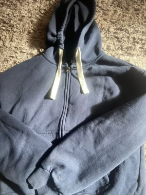 Mörkblå zip hoodie med vita snören - En enkel och stilren mörkblå hoodie med dragkedja framtill och vita dragsnören i huvan. Hoodien har ribbade muddar vid ärmslut och nederkant samt en klassisk känguruficka. Perfekt för en avslappnad look.