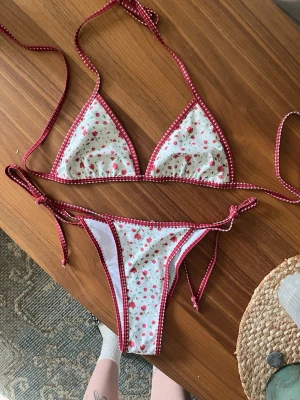 Vit bikini med blommigt mönster - Supersöt bikini med vit botten och små röda blommor. Bikinin har röda kanter med vita prickar och knyt i både topp och trosa för justerbar passform.