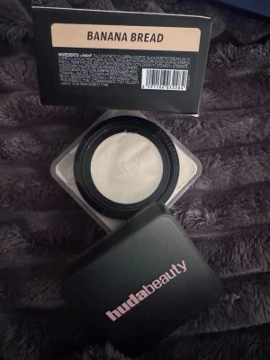 Huda Beauty Easy Bake Banana Bread - Lös baking- och settingpuder från Huda Beauty i nyansen Banana Bread. Kommer i en fyrkantig burk med svart lock och rosa logga. Pudret har en ljus, gulaktig ton som passar för att ljusa upp och fixera makeupen.