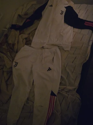 Juventus adidas träningsset vitt - Juventus träningsset från unisport i vitt med svarta och rosa detaljer. Setet består av en zip-jacka med svarta ärmar och rosa ränder samt matchande byxor med Juventus-logga och adidas-märke. Perfekt för fotbollsträning eller chill.