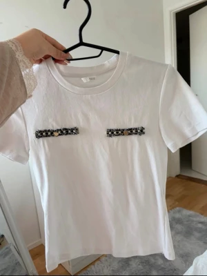 Vit t-shirt med dekorativa detaljer - Vit t-shirt i bomull med korta ärmar och rund hals. Framtill finns två dekorativa svarta och vita band med små guldknappar, vilket ger en unik och trendig look. Perfekt för dig som vill sticka ut lite extra i vardagen.