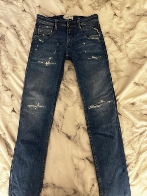 Blå slitna jeans från Replay - Säljer ett par blå jeans från Replay med slitningar och distressed detaljer på både fram- och baksida. Klassisk femficksmodell med smal passform och normal midja. Perfekta för en avslappnad och trendig look.