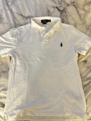 Vit pikétröja från Polo Ralph Lauren - Klassisk vit pikétröja från Polo Ralph Lauren i slim fit-modell. Tröjan har korta ärmar, krage med knappar och den ikoniska broderade loggan på bröstet. Perfekt för en clean och stilren look.
