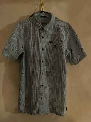 Blå kortärmad vintage skjorta från Columbia - Stilren vintage kortärmad skjorta i ljusblått med smått rutigt mönster från Columbia. Skjortan har klassisk krage, bröstficka och knappar framtill. Storlek står inte tyvärr men jag skulle gissa på M/L vilket motsvarar ungefär 40 i storlek för skjortor