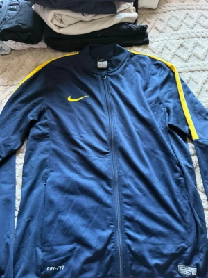 Blå Nike Dri-Fit träningsjacka - Säljer en marinblå Nike Dri-Fit träningsjacka med hel dragkedja och gula detaljer längs axlarna. Jackan har en smal passform och är tillverkad i ett lätt, syntetiskt material som andas. Perfekt för fotbollsträning eller uppvärmning.