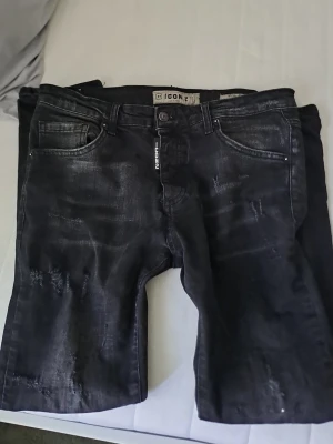 Svarta slitna jeans från ICON2 - Säljer ett par svarta jeans från ICON2 med snygga slitningar och tvättad look. Jeansen har fem fickor, normal midja och raka ben. Perfekta för dig som gillar en trendig och avslappnad stil.