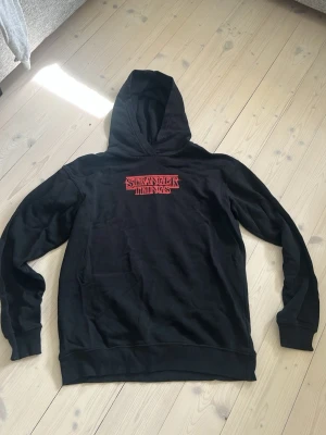 Svart oversize Stranger Things hoodie - Säljer en svart hoodie med Stranger Things-tryck i rött framtill och ett stort färgglatt motiv från serien på ryggen. Hoodien har huva och ribbade muddar vid ärmar och nederkant. Perfekt för dig som älskar serien och vill ha en snygg och bekväm tröja.