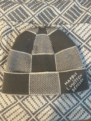 Gråvit rutmönstrad mössa - Säljer en stilren mössa i grått och vitt med rutmönster och texten 'Marque L. Vuitton Déposée' broderad på kanten. Mössan har en modern design och är stickad i mjukt material som passar perfekt för kyliga dagar.