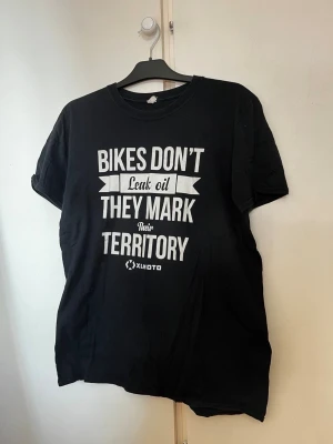 Svart t-shirt med mc texttryck - Svart t-shirt med vitt texttryck: 'Bikes don't leak oil, they mark their territory' och liten XLMOTO-logga. Klassisk rund hals och korta ärmar. Perfekt för dig som gillar motorcyklar och vill ha en chill look.