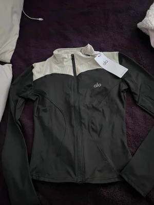 Svart och beige zip-jacka från Alo - Sportig zip-jacka från Alo i svart med beige ribbad överdel och hög krage. Jackan har långa ärmar, figurnära passform och dragkedja framtill. Materialet är en mix av bomull, polyester och elastan för stretch och komfort.