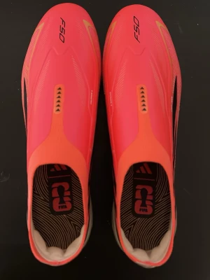 Adidas Hyperfast fotbollsskor - Säljer ett par Adidas Hyperfast fotbollsskor i en riktigt snygg rosa färg med svarta och guldiga detaljer. Skorna har en slimmad, strömlinjeformad design och ovandel i syntetmaterial. Sulan är guldskimrande och har dobbar för bra grepp på planen. Perfekta för dig som vill sticka ut på matchen!