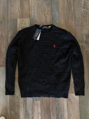 Mörkblå kabelstickad tröja Polo - Snygg mörkblå kabelstickad tröja från Polo Ralph Lauren med rund halsringning och den klassiska röda logotypen broderad på bröstet. Tröjan har långa ärmar och ribbade muddar vid ärmslut och nederkant. Perfekt för lager på lager och stilren look. Tröjan är i storlek M men passar S.