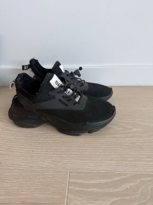 Svarta chunky sneakers med mesh - Säljer ett par svarta chunky sneakers med grov sula och mesh-ovandel. Skorna har svarta snören med snabblås och detaljer i syntetmaterial. Vita logotaggar på plös och häl ger en sportig vibe. Perfekta för dig som gillar streetstyle och komfort.