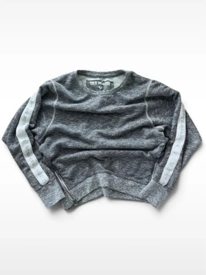 Grå sweatshirt från True Religion - Grå sweatshirt från True Religion med vita band längs ärmarna där märkesnamnet står tryckt. Tröjan har rund halsringning, ribbade muddar och en avslappnad passform. 