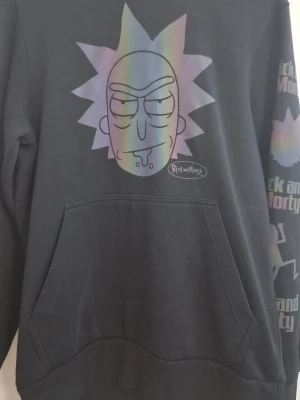 Svart Rick and Morty hoodie - Svart hoodie med stor regnbågsskimrande Rick-tryck på bröstet och Rick and Morty-tryck på ena ärmen. Klassisk känguruficka framtill och huva. Perfekt för dig som gillar serien och vill ha en chill och snygg stil.