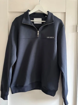 Mörkblå halfzip från Les Deux - Snygg mörkblå sweatshirt med halv dragkedja från Les Deux. Tröjan har broderad logga på bröstet, ribbade muddar och hög krage. Perfekt för en avslappnad men stilren look. Tillverkad i mjukt material som känns skönt mot huden.