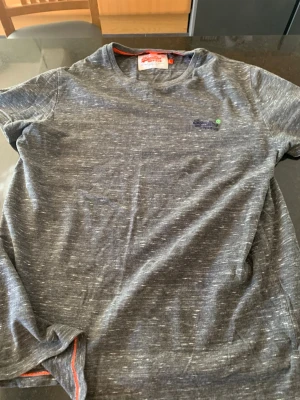 Gråmelerad t-shirt från Superdry - Säljer en gråmelerad t-shirt från Superdry med broderad logga på bröstet. T-shirten har rund halsringning och korta ärmar. Skön och avslappnad passform, perfekt till jeans eller shorts.