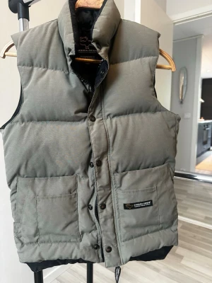 Kakifärgad dunväst från Canada Goose - Snygg dunväst från Canada Goose med två stora framfickor. Fick skick förutom att det är lite skavt på vissa svarta knappar vid tvätt. Storlek 2xs 