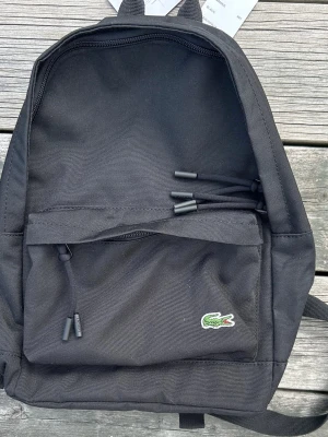Svart ryggsäck från Lacoste - Snygg svart ryggsäck från Lacoste i slitstark polyester. Klassisk design med dragkedja, framficka och det ikoniska gröna krokodilmärket på framsidan. Perfekt för skola eller cityhäng med justerbara axelband och praktiskt bärhandtag.Helt ny och kommer med originaltaggen kvar.