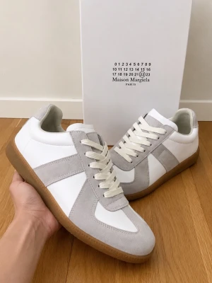Maison Margiela vita och grå sneakers - Maison Margiela sneakers i vitt och ljusgrått med ovandel i läder och mocka. Klassisk låg modell med vita snören och gummisula. Snygg minimalistisk design och diskret logga på plösen. Perfekta för dig som gillar stilrena och trendiga skor.