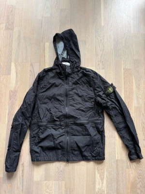 Svart vindjacka från Stone Island - Snygg svart vindjacka från Stone Island med huva och dragkedja framtill. Jackan har den klassiska Stone Island-loggan på vänster ärm och två fickor med dragkedja. 