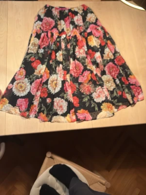 Blommig långkjol från Camilla Thulin oanvänd - Säljer en långkjol från Camilla Thulin med svart botten och stora blommor i rosa, rött, orange och vitt. Kjolen har ett flowy fall och resår i midjan för skön passform. Perfekt för dig som vill sticka ut med färg och mönster!