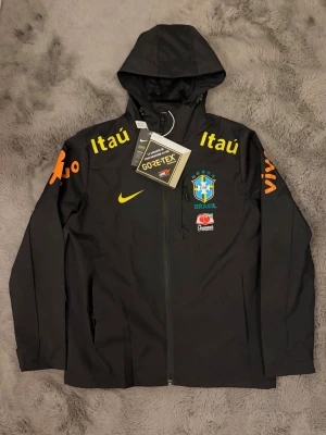 Svart Brasil Nike vindjacka med huva - Svart vindjacka från Nike med Brasil-logga och sponsormärken som Itaú och Guaraná Antarctica. Jackan har huva, hel dragkedja och är tillverkad i GORE-TEX-material. Färgdetaljer i gult och orange på bröst och ärmar. Perfekt för dig som gillar sportig stil.