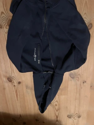 Marinblå hoodie från Sail Racing - Säljer en marinblå hoodie från Sail Racing med dragkedja framtill och en bröstficka med dragkedja. Hoodien har justerbar huva med snören och är tillverkad i mjukt material. Perfekt för dig som gillar sportig och stilren look.