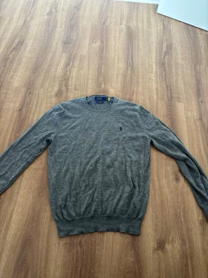 Grå stickad tröja Polo Ralph Lauren - Snygg grå stickad tröja från Polo Ralph Lauren i slim fit-modell. Tillverkad av tvättbar merinoull och har rund halsringning samt diskret logga broderad på bröstet. Perfekt för lager på lager och enkel att matcha med det mesta. Pris är diskuterbart vid snabb affär. Passar även bra om man har storlek M.