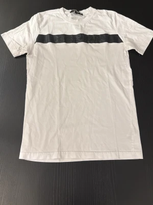 Vit t-shirt från Dolce & Gabbana - Vit t-shirt från Dolce & Gabbana med svart band och logga över bröstet. Klassisk passform och rund halsringning. Tillverkad i mjuk bomull och har korta ärmar. Perfekt för en stilren och enkel look.