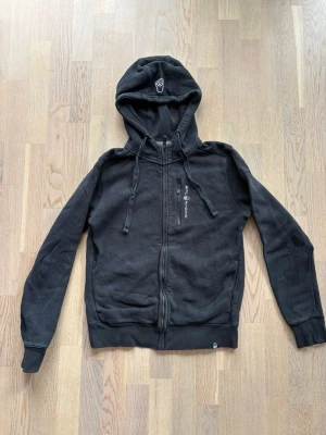 Svart hoodie från Sail Racing - Svart hoodie med dragkedja från Sail Racing. Har broderad logga på bröstet och stor Sail Racing-text på ryggen. Använd men mycket bra skick