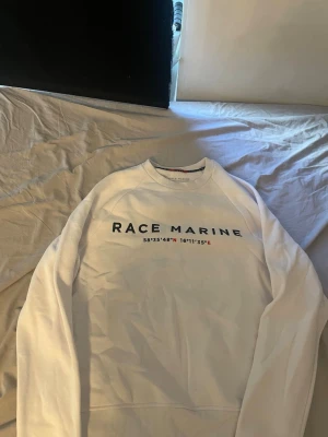 Vit sweatshirt från Race Marine - Vit sweatshirt från Race Marine med tryckt logga och koordinater på bröstet. Tröjan har rund halsringning, ribbade muddar och är tillverkad i mjukt material. Perfekt för en clean och sportig stil.