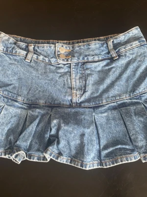 Blå plisserad y2k jeanskjol - Snygg blå jeanskjol med plisserad nederdel och dubbla knappar framtill. Kjolen har bälteshällor och klassisk femficksdesign. Perfekt för dig som vill ha en y2k look.