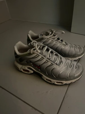 Nike Air Max Plus vita och grå sneakers - Nike Air Max Plus sneakers i vitt och grått med röda detaljer. Skorna har en vågig design på ovandelen, synlig Air-dämpning i sulan och svart yttersula. Tillverkade i textil och syntetmaterial med klassisk snörning och låg profil.
