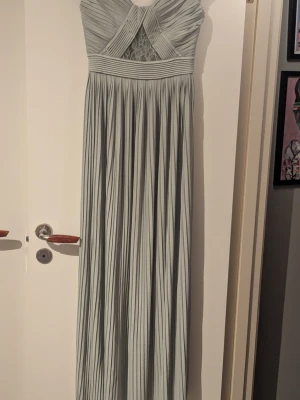 Ljusgrön långklänning med spetsdetalj - Elegant ljusgrön långklänning med plisserad kjol och omlott överdel. Klänningen har spetsdetalj framtill och bred midjeresår. Offshoulder-modell med fina veck och mjukt fallande tyg, perfekt för speciella tillfällen. 