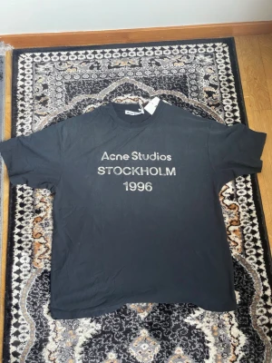 Svart Acne Studios t-shirt Stockholm - Svart t-shirt från Acne Studios med trycket 'Acne Studios STOCKHOLM 1996' på bröstet. Klassisk passform med rund hals och korta ärmar. Tillverkad i mjuk bomull för en bekväm känsla.