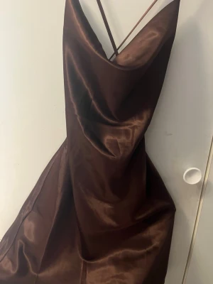 Brun satin slipklänning med smala band - Snygg brun långklänning i satin med tunna axelband som korsas i ryggen. Klänningen har en mjukt draperad urringning och glansigt material som ger en lyxig känsla. Perfekt för dig som vill ha en elegant och trendig look. Skulle säga att den passar XS eller S