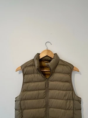 Olivgrön dunväst från Uniqlo - Snygg olivgrön dunväst från Uniqlo i ultralätt material. Västen har hög krage, dragkedja framtill och quiltad design. Perfekt för lager-på-lager och enkel att matcha med olika outfits.