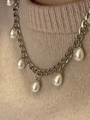 Kedjehalsband med droppformade pärlor - Snyggt halsband med grov silverfärgad kedja och sju droppformade vita pärlor som hänger längs kedjan. Perfekt för att ge en cool och trendig touch till din outfit. Materialet är rostfritt och pärlorna.