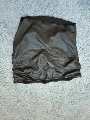 Svart kortkjol i skinnimitation - Säljer en svart kortkjol i skinnimitation med snygga rynkade detaljer på sidorna. Kjolen har en tight passform och är perfekt för dig som gillar en edgy stil. Enkel att matcha med olika toppar och accessoarer.