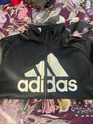 Svart Adidas ziptröja med logga - Säljer en svart ziptröja från Adidas med stor vit logga och text på framsidan. Tröjan har hög krage och dragkedja framtill. Perfekt för träning eller chill. Materialet är mjukt och stretchigt för skön passform.