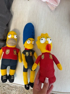 Simpsons mjukisdjur i fotbollströjor - Tre mjukisdjur föreställande Homer, Marge och Lisa från Simpsons, klädda i spanska landslagets fotbollströjor. Homer och Lisa har röda tröjor, Marge har svart tröja och blått hår. Alla har broderade detaljer och är gjorda i mjukt tyg.