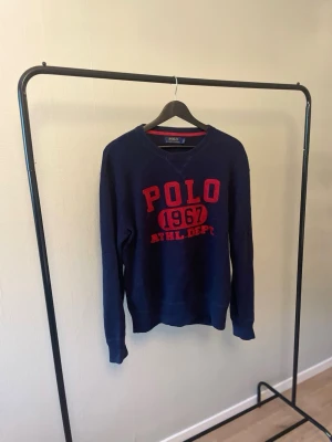 Polo Ralph Lauren Sweatshirt👕 - Polo Ralph Lauren sweatshirt – Strl S 🔥 Mörkblå sweatshirt från Polo Ralph Lauren med klassiskt "POLO 1967 ATHL. DEPT."-tryck i rött. Stilren och tidlös modell i fint begagnat skick – perfekt till både jeans och chinos. 📏 Storlek: S 📦 Skick: Bra begagnat skick 💬 Hör gärna av dig vid frågor eller om du vill ha fler bilder!