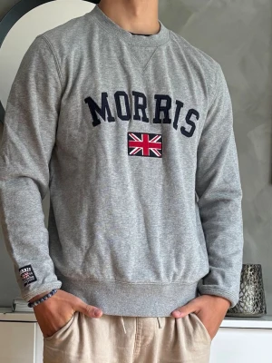 Grå sweatshirt från Morris - Grå sweatshirt från Morris med mörkblå text och brittisk flagga broderad på bröstet. Tröjan har rund halsringning, ribbade muddar vid ärmslut och nederkant samt en liten patch på ärmen. Perfekt för en avslappnad och stilren look.