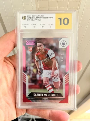 Gabriel Martinelli Pink Lava #198 kort - Säljer ett samlarkort från 2021-22 Score P/L, Gabriel Martinelli Pink Lava 8/25, graderat 10 av PCG. Kortet har rosa och röda detaljer, visar Martinelli i Arsenal-tröja och är inplastat i hårt skydd. Perfekt för fotbollssamlare!