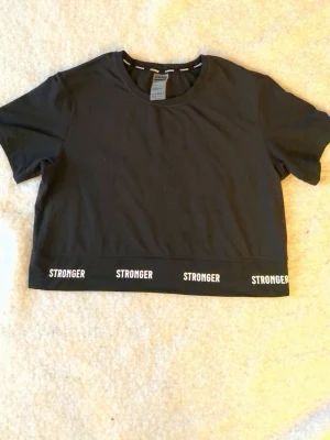 Svart crop top från Stronger - Snygg svart crop top från Stronger med korta ärmar och rund halsringning. Nedtill finns en bred resår med loggan 'STRONGER' tryckt flera gånger i vitt. Perfekt till träning eller en sportig look.