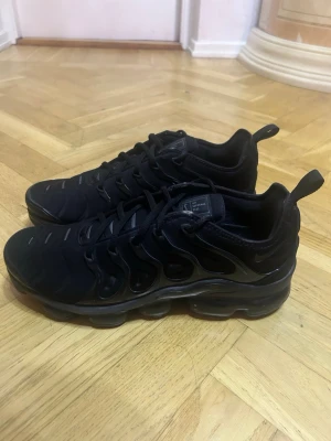 Svarta Nike Air VaporMax Plus - Säljer ett par helsvarta Nike Air VaporMax Plus sneakers med ikonisk bubbelsula och detaljerad ovandel i syntetmaterial. Skorna har dragflik bak, snörning och diskreta loggor. Perfekta för dig som gillar sportig och modern stil.