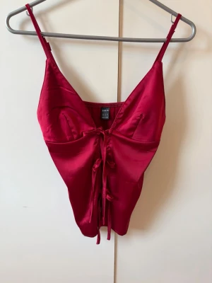 Röd satinbody med snörning från SHEIN - Snygg röd body i glansigt satinmaterial från SHEIN. Modellen har tunna axelband, djup urringning och dekorativ snörning framtill. Perfekt för dig som vill ha något extra till underkläderna. Materialet känns mjukt och följsamt mot huden.