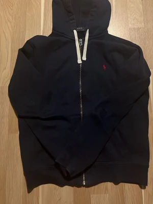 Marinblå hoodie från Polo  Ralph Lauren  - Snygg marinblå hoodie från Polo Ralph Lauren med dragkedja framtill och vit dragsko i huvan. Klassisk röd logga broderad på bröstet. Tillverkad i mjukt material som passar perfekt för en avslappnad stil.