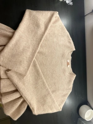 Beige stickad tröja med volang - Supersöt beige stickad tröja med rund halsringning och långa ärmar. Tröjan har en unik volangdetalj nedtill som ger en trendig och cool look. Perfekt att styla med jeans eller kjol för en snygg outfit.